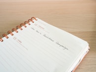 An open spiral-bound planner lies on a wooden surface. The pages are dated December 12th and 13th, with handwritten text 'New Year's Resolutions Campaign' on the 12th. The planner is designed with lines and dates printed in orange.