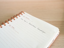 An open spiral-bound planner lies on a wooden surface. The pages are dated December 12th and 13th, with handwritten text 'New Year's Resolutions Campaign' on the 12th. The planner is designed with lines and dates printed in orange.