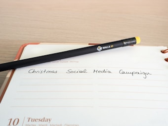 A black pencil with a yellow eraser lies diagonally across an open planner, with the words 'Christmas Social Media Campaign' written on one of the lined pages. The date 'Tuesday 10' is visible at the bottom of the page.