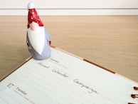 A small gnome figurine with a red hat stands on an open planner. The planner displays the text 'Advent Calendar Campaign' and shows Tuesday the 17th.