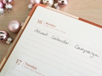 A planner or calendar is open to Monday, the 16th, with the words 'Advent Calendar Campaign' written in black ink. Surrounding the planner are shiny, metallic Christmas ornaments in shades of pink and silver. The planner has a wooden table underneath.