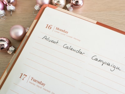 A planner or calendar is open to Monday, the 16th, with the words 'Advent Calendar Campaign' written in black ink. Surrounding the planner are shiny, metallic Christmas ornaments in shades of pink and silver. The planner has a wooden table underneath.