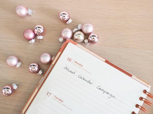 A calendar opened to the 16th of the month with the text 'Advent Calendar Campaign' written on it, placed on a light wooden surface. Surrounding the calendar are several pinkish and metallic small Christmas ornaments.