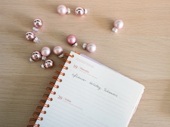 A spiral-bound notebook is open on a wooden surface, displaying a page with handwritten text that reads 'Influencer Holiday Takeovers.' Pink ornaments, resembling Christmas baubles, are scattered around the notebook, creating a festive atmosphere.