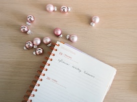 A spiral-bound notebook is open on a wooden surface, displaying a page with handwritten text that reads 'Influencer Holiday Takeovers.' Pink ornaments, resembling Christmas baubles, are scattered around the notebook, creating a festive atmosphere.