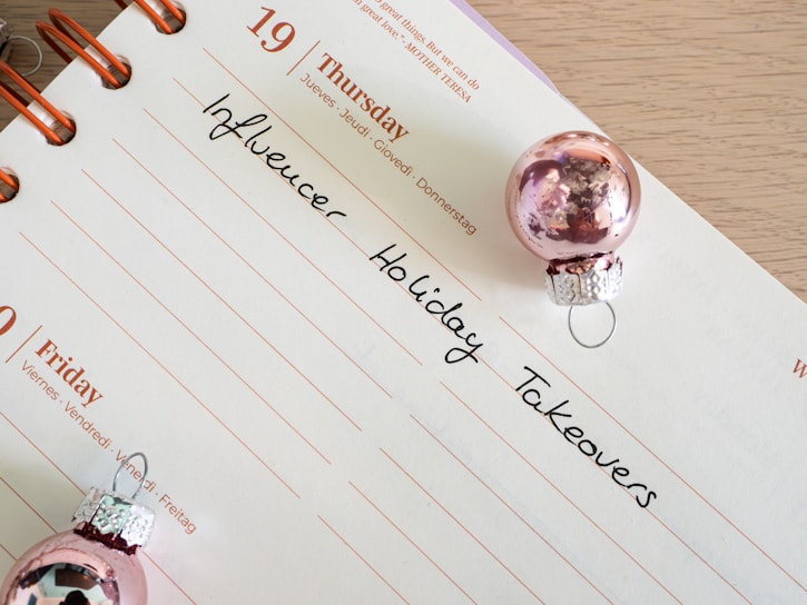 A calendar page open to Thursday the 19th with handwritten notes reading 'Influencer Holiday Takeovers.' The page is decorated with two small pink Christmas ornaments.