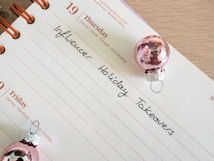 A calendar page open to Thursday the 19th with handwritten notes reading 'Influencer Holiday Takeovers.' The page is decorated with two small pink Christmas ornaments.