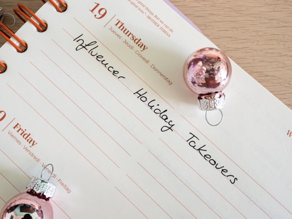 A calendar page open to Thursday the 19th with handwritten notes reading 'Influencer Holiday Takeovers.' The page is decorated with two small pink Christmas ornaments.