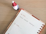 A tabletop scene featuring an open planner with copper rings displaying a week that includes Monday, the 16th, and Tuesday, the 17th. A note on the Monday page reads 'Advent Calendar Campaign.' To the top left of the planner is a small figurine of a Santa Claus or gnome, wearing a red hat with white spots and a red coat.