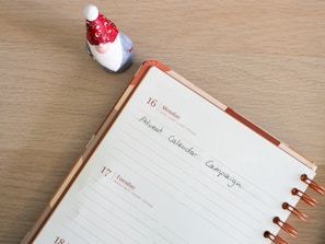 A tabletop scene featuring an open planner with copper rings displaying a week that includes Monday, the 16th, and Tuesday, the 17th. A note on the Monday page reads 'Advent Calendar Campaign.' To the top left of the planner is a small figurine of a Santa Claus or gnome, wearing a red hat with white spots and a red coat.