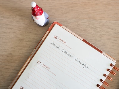 A tabletop scene featuring an open planner with copper rings displaying a week that includes Monday, the 16th, and Tuesday, the 17th. A note on the Monday page reads 'Advent Calendar Campaign.' To the top left of the planner is a small figurine of a Santa Claus or gnome, wearing a red hat with white spots and a red coat.