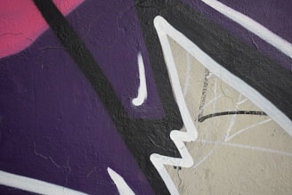 Abstract street art featuring bold, angular shapes and overlapping lines. The composition consists of dark purple, pink, and beige backgrounds, accentuated by black and white lines.