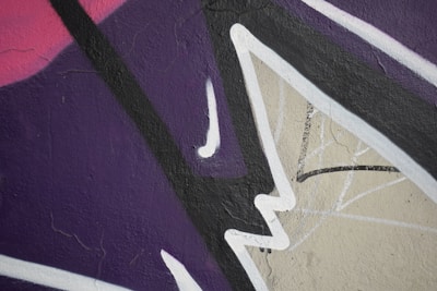 Abstract street art featuring bold, angular shapes and overlapping lines. The composition consists of dark purple, pink, and beige backgrounds, accentuated by black and white lines.