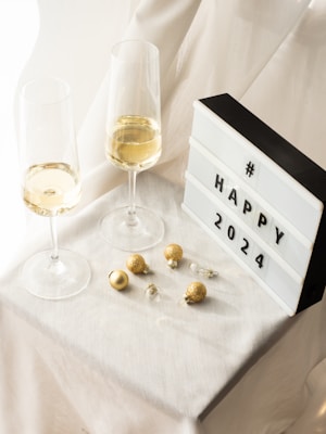 Two champagne glasses filled with a sparkling beverage are placed on a white cloth-covered table, accompanied by a light box displaying the message '# HAPPY 2024'. Golden and clear baubles are scattered on the table, adding a festive touch.