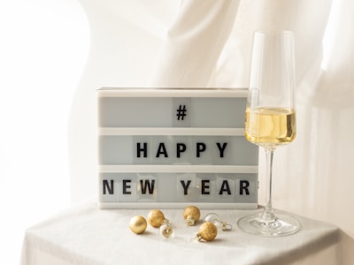 A lightbox displays the message 'Happy New Year' alongside a glass of champagne. A few small golden ornaments are scattered nearby on a white surface. The scene is warmly lit, suggesting a celebratory and festive atmosphere.