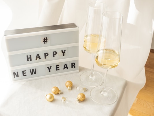 A lightbox displaying the message '# HAPPY NEW YEAR' is placed on a white surface. To the right of the lightbox, two tall champagne glasses are filled with a light-colored beverage. Small, gold-colored decorative baubles are scattered on the surface next to the glasses. The scene is brightly lit with soft shadows.