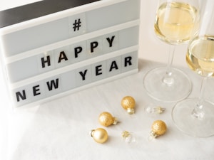 A lightbox sign displays the message HAPPY NEW YEAR, accompanied by two glasses of champagne and several small, golden Christmas ornaments on a white tablecloth.