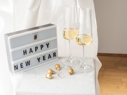 A lightbox displays the message 'HAPPY NEW YEAR' in bold black letters. Beside it, two champagne flutes contain a golden bubbly drink, elegantly placed on a white surface. Several small, gold and white Christmas ornaments are scattered nearby, creating a festive atmosphere against a soft, flowing white curtain backdrop.