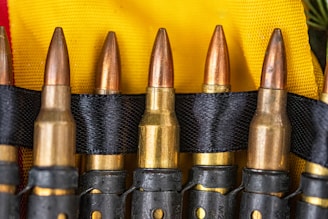 Several bullets are neatly aligned in a black belt against a background of yellow fabric. The bullets have a brass casing with copper-colored tips, and the black belt holds them securely in place.