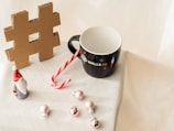 A black mug with 'WALLS.IO' printed on it sits on a white surface. Next to it is a red and white candy cane, a small ceramic figure of a Santa-like gnome, a cardboard cutout of a hashtag symbol, and several small pink Christmas ornaments. The setting suggests a festive, holiday theme.