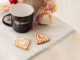 A black mug with a logo sits next to a fuzzy teddy bear holding a candy cane. Two heart-shaped cookies with white icing detail rest on a white tablecloth.