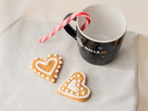 On a light-colored fabric surface rests a black mug with a white interior, featuring a logo and the text 'Walls.io' on its side. A candy cane is placed inside the mug, with its red and white striped body leaning out. Two heart-shaped cookies are positioned in front of the mug, decorated with white icing, creating a pleasant arrangement.