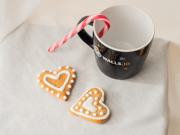 On a light-colored fabric surface rests a black mug with a white interior, featuring a logo and the text 'Walls.io' on its side. A candy cane is placed inside the mug, with its red and white striped body leaning out. Two heart-shaped cookies are positioned in front of the mug, decorated with white icing, creating a pleasant arrangement.