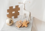 A decorative scene features a large gold hashtag symbol, a white ceramic deer figurine, a gold ornament ball, and a plate of gingerbread cookies shaped like a house and a person. The arrangement is set on a white cloth and conveys a festive, minimalistic aesthetic.