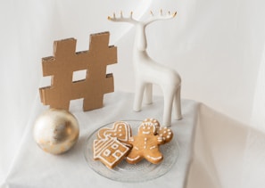 A decorative scene features a large gold hashtag symbol, a white ceramic deer figurine, a gold ornament ball, and a plate of gingerbread cookies shaped like a house and a person. The arrangement is set on a white cloth and conveys a festive, minimalistic aesthetic.