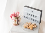 A holiday-themed display features a decorative mug filled with peppermint candy canes alongside a lightbox sign displaying '# HAPPY HOLIDAYS'. Next to them, a clear glass plate holds three gingerbread cookies shaped like a house, a heart, and a person. The background consists of soft, white fabric, creating a festive and cozy atmosphere.
