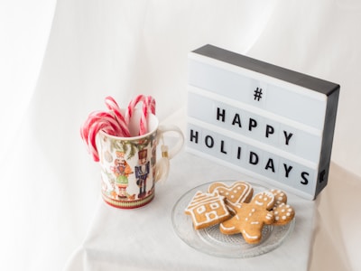 A holiday-themed display features a decorative mug filled with peppermint candy canes alongside a lightbox sign displaying '# HAPPY HOLIDAYS'. Next to them, a clear glass plate holds three gingerbread cookies shaped like a house, a heart, and a person. The background consists of soft, white fabric, creating a festive and cozy atmosphere.