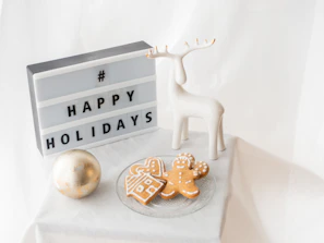 A lightbox sign displaying the message '# HAPPY HOLIDAYS' sits next to a white ceramic reindeer. A clear glass plate holds two gingerbread cookies shaped like a house and a person, decorated with white icing. A large, silver and gold decorative bauble is positioned to the left, all set against a soft white fabric background.