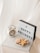 A lightbox with the text 'Happy Holidays' is positioned on a white surface alongside a vintage alarm clock featuring a Santa Claus image. In front of them is a clear glass plate holding several gingerbread cookies shaped like a gingerbread man, a house, and a heart.