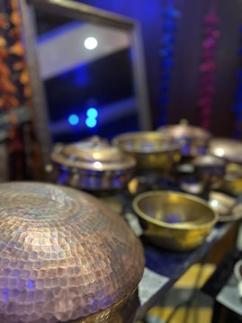 Various brass or copper bowls with a hammered texture are arranged on a surface. The background features dim lighting with blue and purple hues, contributing to a reflective and warm ambience. A mirror partially visible in the background reflects some light.