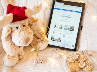 A plush reindeer toy wearing a red hat sits next to a tablet displaying a webpage with the hashtag #LoveWallsio. The scene is decorated with string lights, metallic baubles, and a plate of gingerbread cookies shaped like a house, tree, and gingerbread person, all set on a knitted white background.