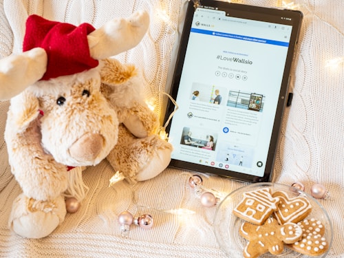 A plush reindeer toy wearing a red hat sits next to a tablet displaying a webpage with the hashtag #LoveWallsio. The scene is decorated with string lights, metallic baubles, and a plate of gingerbread cookies shaped like a house, tree, and gingerbread person, all set on a knitted white background.