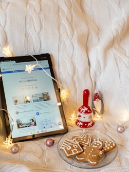 A cozy scene features a tablet displaying a web page with the hashtag #LoveWallsio, placed on a textured white cable-knit blanket. Festive string lights with star-shaped bulbs surround the tablet, adding a warm glow. Next to the tablet is a red decorative bell with white patterns of reindeer and snowflakes. Nearby, a small red and white gnome figurine is visible. In front of the tablet, a glass plate holds gingerbread cookies shaped like a house, a heart, and a gingerbread man. Several pastel ornaments are scattered across the blanket.