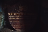 The image depicts an old, dark, and weathered tombstone with worn-out inscriptions. The faint lighting casts shadows, making it challenging to read the entire text. The tombstone appears to be made of stone or marble and is found in a presumably damp and aged environment.