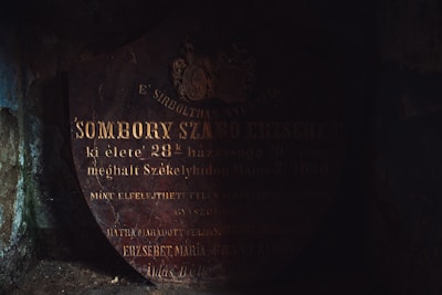 The image depicts an old, dark, and weathered tombstone with worn-out inscriptions. The faint lighting casts shadows, making it challenging to read the entire text. The tombstone appears to be made of stone or marble and is found in a presumably damp and aged environment.