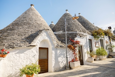 Traditional stone houses with conical roofs are set against a clear blue sky. These structures are painted white with textured stone rooftops. Climbing plants and potted flowers decorate the exterior, adding a touch of greenery. A single street lamp stands in front of one of the entrances.