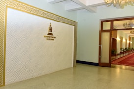 An interior hallway featuring a decorative white wall with geometric patterns bordered in gold, displaying a crest and text emblem. The adjacent area opens into a corridor adorned with a red carpet, potted plants, and chandeliers hanging from the ceiling.