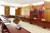 A large, formal conference room with a U-shaped wooden table surrounded by chairs with tan cushions. Several identical flags with a red and blue design are displayed along one wall, and a large emblem with a circular motif is mounted above them. A plant is positioned on the table as a centerpiece. The room is well-lit with natural light from large windows with yellow curtains and artificial light from a ceiling chandelier.