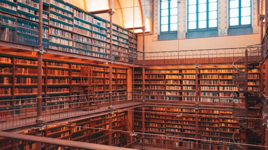 A spacious library filled with numerous books organized on tall shelves that reach up to a high ceiling. The interior is structured with multiple levels, connected by a spiral staircase. Large windows allow natural light to illuminate the warm wooden tones of the shelves and floor.