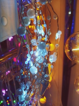 A close-up view of a decorative string of lights with spherical ornaments. The ornaments are multi-colored and translucent, with glowing blue and white lights encased within them. The background is blurred but contains more lights that are predominantly yellow, purple, and blue, adding a festive ambiance to the scene.