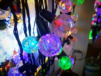 A close-up view of colorful, round light bulbs strung together with dark cords, emitting vibrant hues of blue, purple, green, and orange. The bulbs have a textured, sparkling surface, enhancing their glow. The background is softly blurred with additional lights and colors visible.