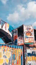Shipping containers are creatively adorned with vibrant graffiti art. The vivid colors and abstract designs cover the surfaces, adding a splash of urban art to the industrial structures. The blue sky with scattered clouds forms the backdrop.