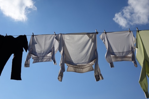 A clear sky serves as the backdrop for several pieces of clothing hanging on a drying line, including white shirts and other garments. The bright sunlight creates shadows and highlights the texture of the fabric.