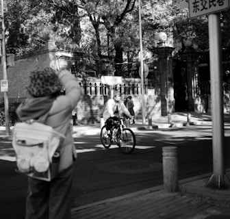 A black and white urban scene depicting a cyclist riding down a street. In the foreground, a person with a backpack and hat appears to be taking a photo or observing the scene. Trees and ornate brick architecture form the background, with sunlight casting shadows on the street and buildings.