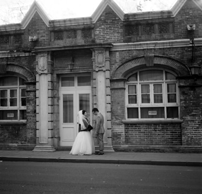 A couple dressed in wedding attire stands in front of an old brick building. The bride is wearing a veil and a white gown, while the groom is in a suit. They are positioned on a sidewalk in front of a weathered structure with arched windows and decorative stone elements.