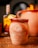 A rustic clay cup with the word 'Mezcally' embossed on its surface, garnished with slices of lime and other fruit on the rim, placed on a wooden table. In the blurred background, there is a bottle and another clay pot, creating a warm and inviting atmosphere.